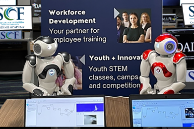 NAO robots sitting on a table in front of a banner with computer screens in the background